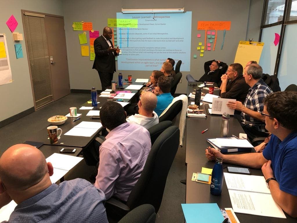 An instructor explaining Agile retrospectives to a group, with the team's own exercise boards and sticky notes covering the wall beside the slide.