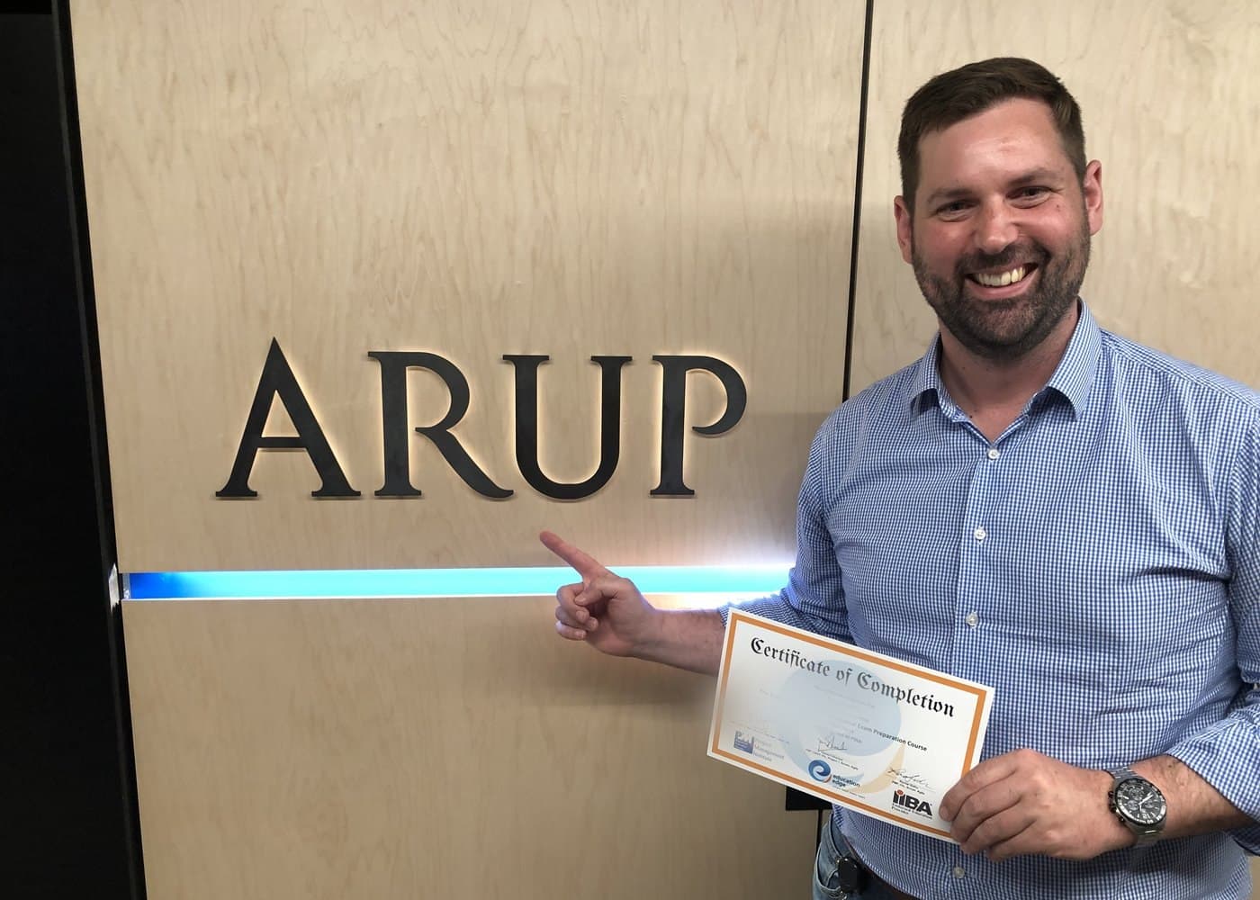 A participant holding an Education Edge Certificate of Completion for the Project Management Professional Exam Preparation Course, beside the ARUP sign at the firm's entrance.