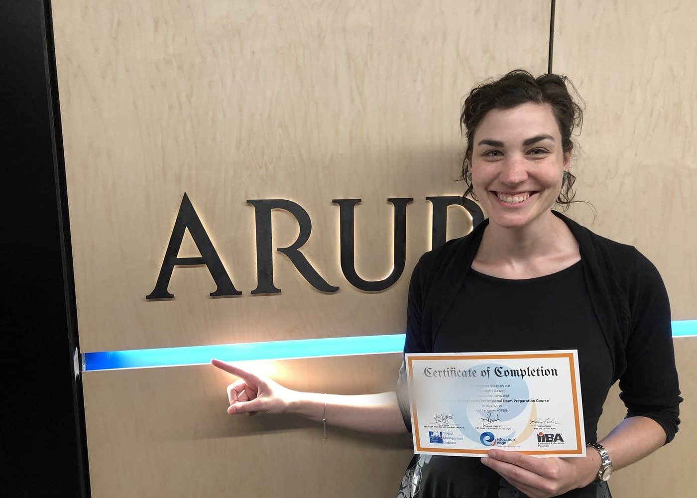 A course participant holding a certificate of completion beside the ARUP sign in the company's office.