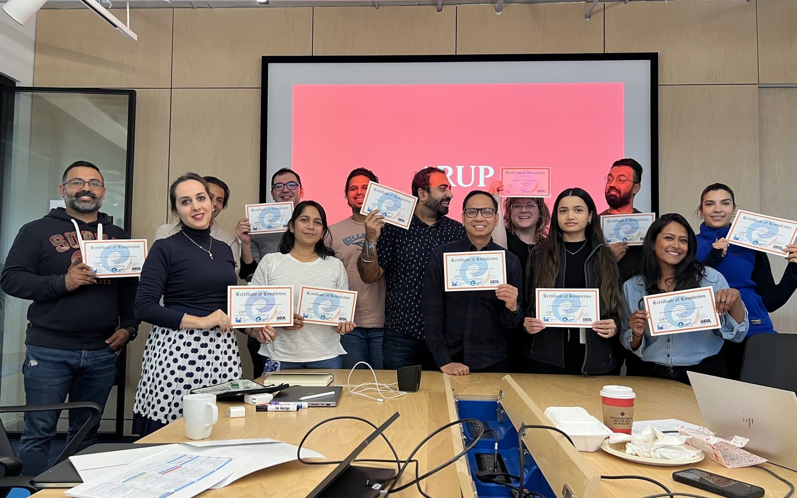 An ARUP cohort standing together at the end of their course, each holding one of our certificates of completion, the ARUP mark on the screen behind them.