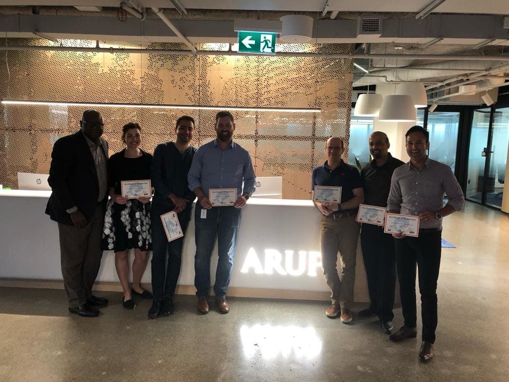 Seven course participants holding certificates of completion at the ARUP office reception desk.
