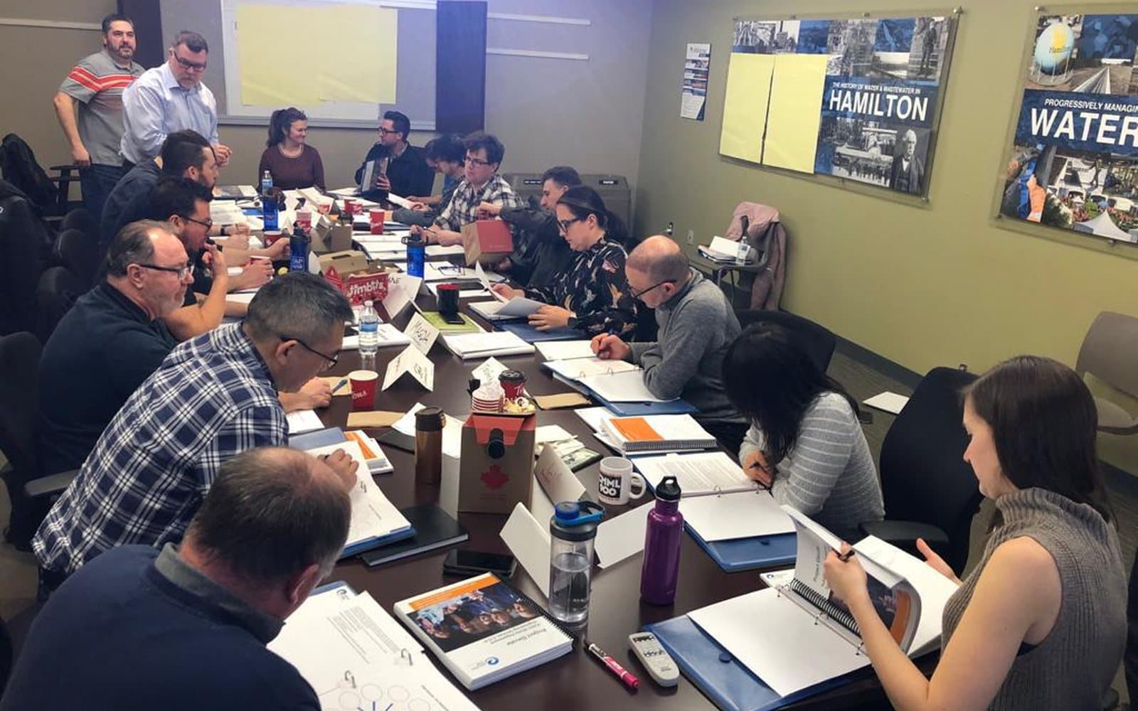 Workshop participants writing in their course workbooks along a long boardroom table, with two facilitators standing at the front of the room.