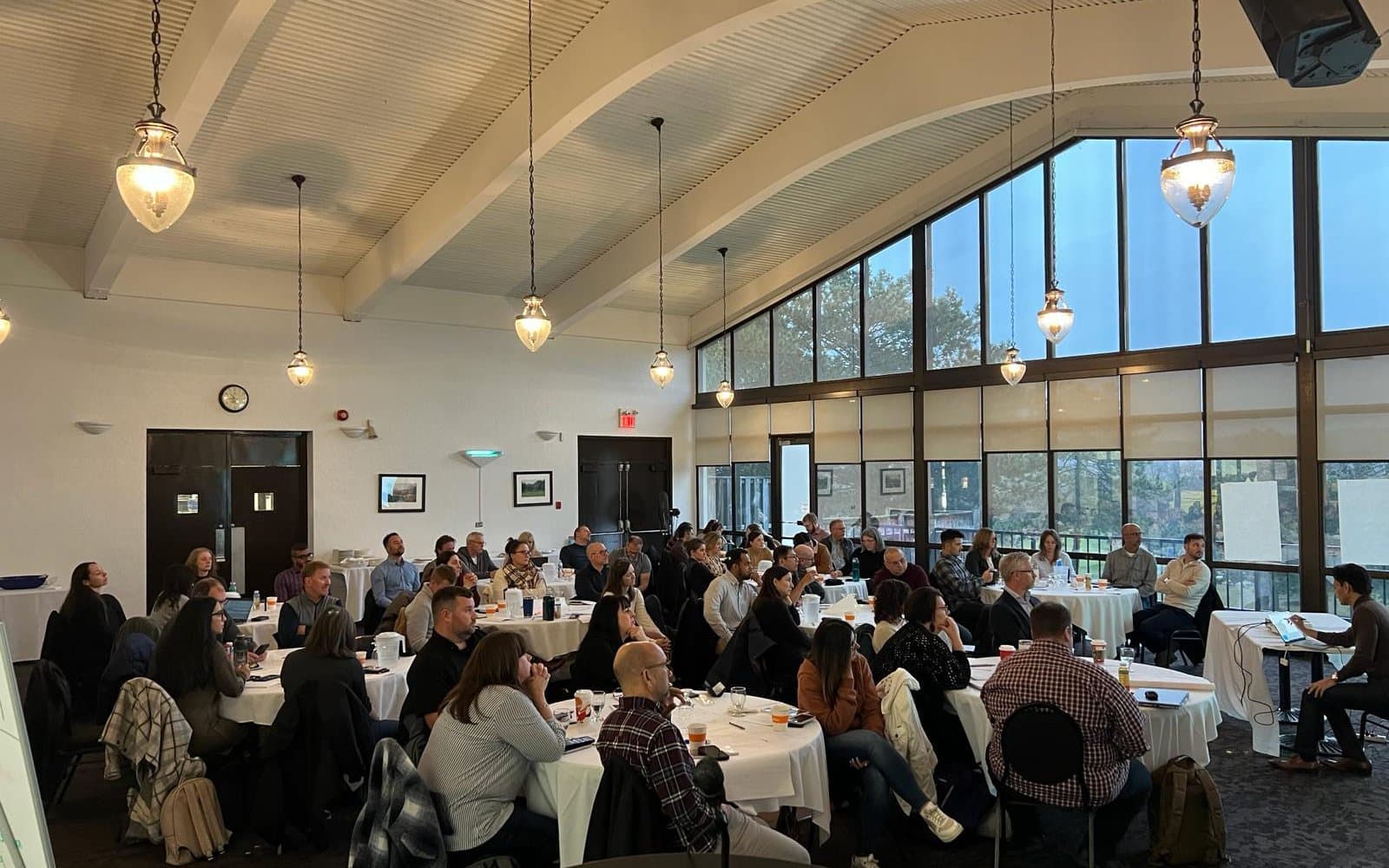 A full room of participants seated at round tables under a vaulted ceiling and a wall of tall windows, with an instructor working at a laptop at a table to one side.