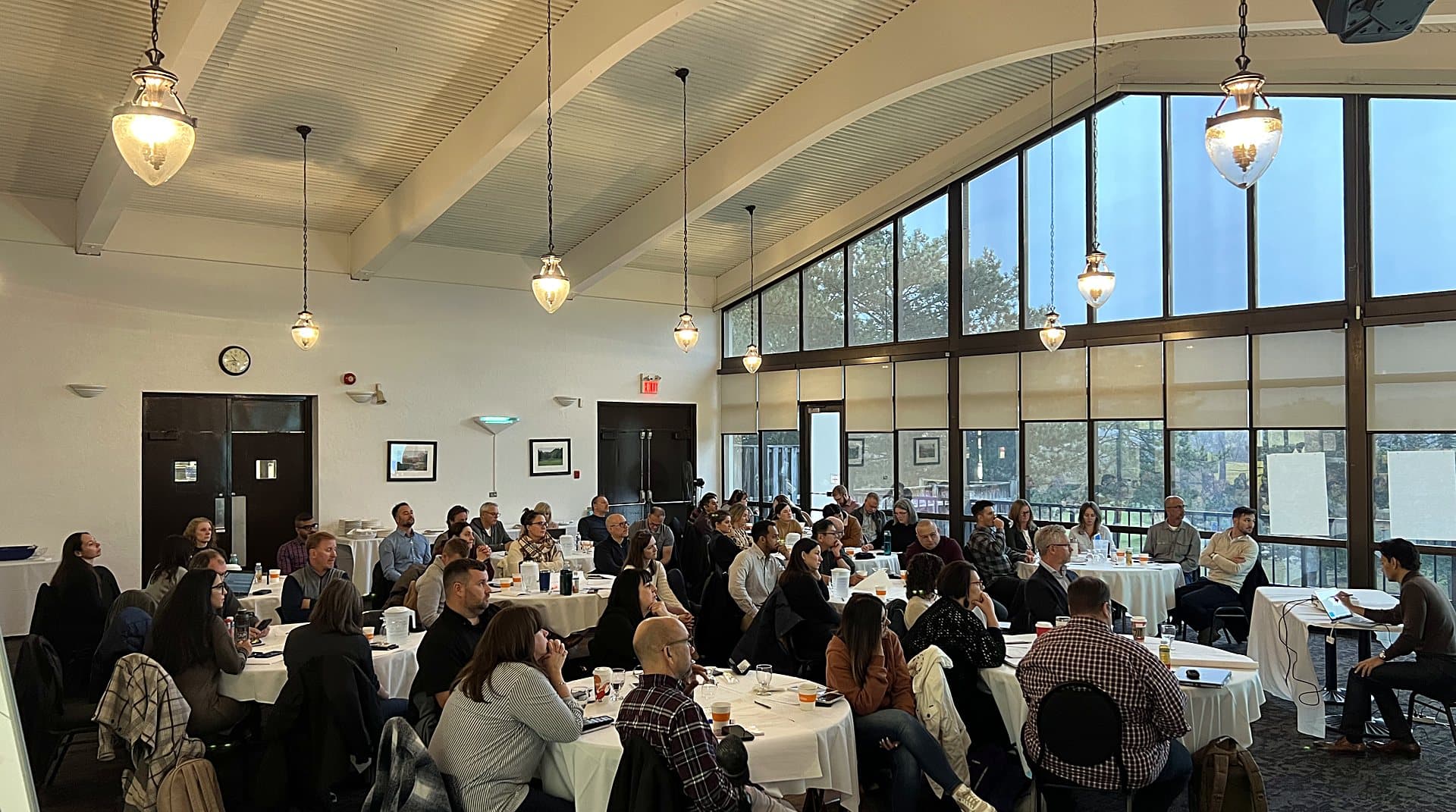 A hall of about fifty people seated at round tables under a vaulted ceiling and a tall wall of windows, facing a presenter at a laptop on the far side of the room.