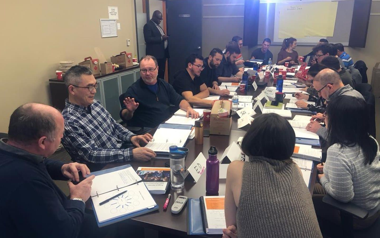 A boardroom cohort working through course binders around a long table, with the trainer standing at the door.