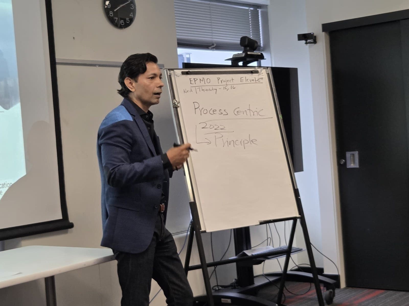 Hemant Dhariyal in a blue blazer, marker in hand, mid-sentence beside a flipchart he has written EPMO Project Elevate, Process Centric and 2022 Principle on, in a meeting room with a wall clock and a projector screen.