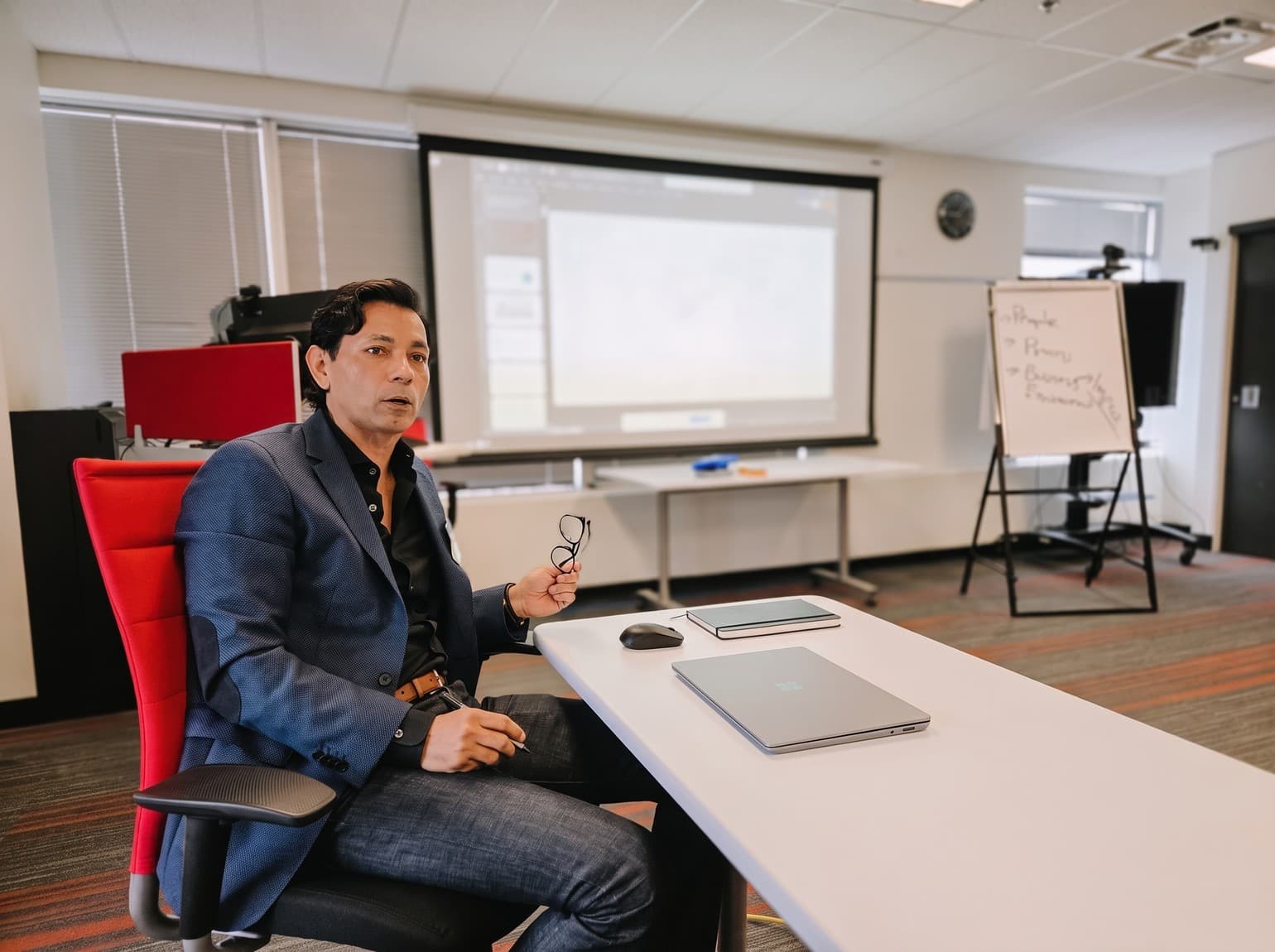 Hemant Dhariyal seated at a training table mid-sentence, glasses in hand, a laptop open beside him and a projector screen behind.
